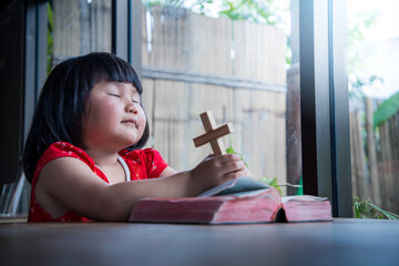 Little girl praying and holding wooden cross on bible at home, child's pure faith