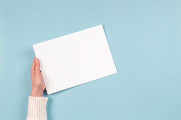 Female hands holding blank white paper sheet on light blue background. Top view. Mockup paper with...