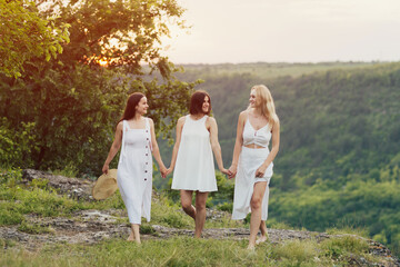 Naklejka na ściany i meble Company of three young female friends having fun, enjoy green hill landscape and beautiful sunset at summer picnic.