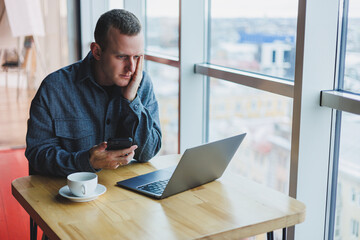 A young handsome man sits at a table and looks with surprise at the screen of his laptop, remote work