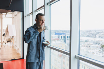 Fototapeta premium A young man of European appearance in a casual suit drinks coffee and looks out the window