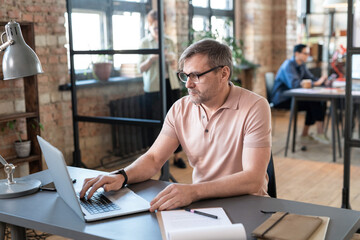 Fototapeta premium Mature businessman in eyeglasses sitting at the table and typing on laptop, he doing online work at office