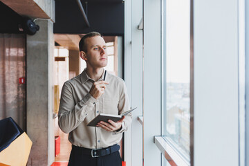 A young businessman stands at a panoramic window in a skyscraper and makes notes in his notebook with a pen. A man in a shirt and trousers of European appearance