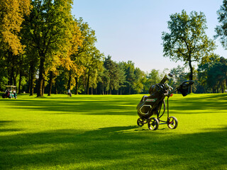 The golfer walks along the fairway at a golf course in the summer on a sunny day. In the foreground, a cart parked on the fairway, with a bag full of golf clubs.