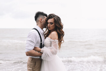 The bride and groom in wedding dresses hug and walk together along the sea in nature on an outdoor trip. Husband and wife in love spend their honeymoon. Selective focus