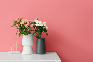 Vases with beautiful roses and decor on table near pink wall