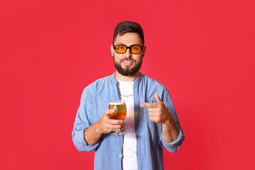 Handsome man in sunglasses pointing at glass of beer on red background