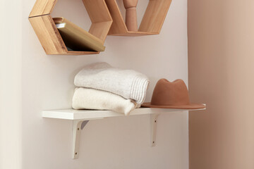 Shelf with stack of warm sweaters and hat hanging on light wall