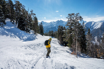Beautiful landscape of the Arkhyz ski resort with mountains, snow, forest and man snowboarder in yellow jacket and backpack on a sunny winter day. Caucasus  Mountains, Russia