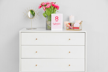 Greeting card with text HAPPY WOMEN'S DAY, tulips and decorative cosmetics on chest of drawers near light wall
