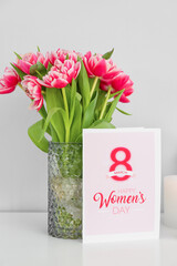 Greeting card with text HAPPY WOMEN'S DAY and vase with tulips on table near light wall