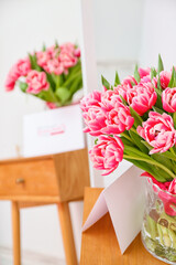 Vase with beautiful tulips and greeting card on wooden table near light wall. International Women's Day celebration