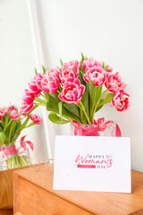 Greeting card with text HAPPY WOMAN'S DAY and bouquet of tulips on wooden table near light wall