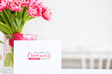 Vase with bouquet of beautiful tulips and greeting card with text HAPPY WOMAN'S DAY on table, closeup