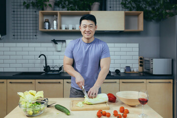 The man is at home alone, preparing a salad, slicing vegetables in the kitchen, the Asian looks at the camera and smiles