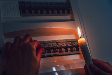 Energy crisis. Hand in complete darkness holding a candle to investigate a home fuse box during a...