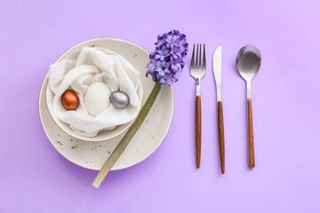 Table setting with Easter eggs and flowers on lilac background