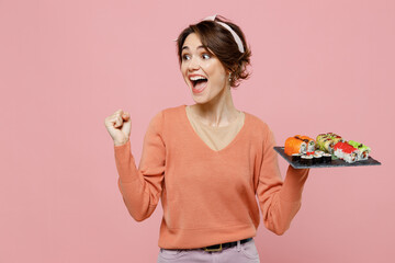Young woman in sweater hold makizushi sushi roll served on black plate traditional japanese food do winner gesture celebrate clench fists say yes look aside isolated on plain pastel pink background