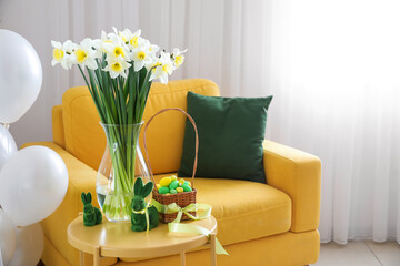 Vase with flowers, Easter bunnies and eggs on table near armchair in room