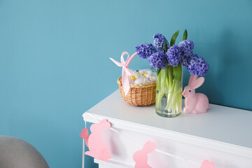 Vase with beautiful flowers, Easter eggs in basket and bunny on mantelpiece near color wall