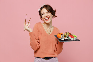 Young smiling happy cheerful cool woman 20s in casual clothes hold in hand makizushi sushi roll served on black plate traditional japanese food chopsticks isolated on plain pastel pink background