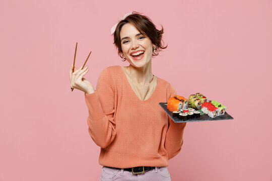 Young smiling happy cheerful cool woman 20s in casual clothes hold in hand makizushi sushi roll served on black plate traditional japanese food chopsticks isolated on plain pastel pink background