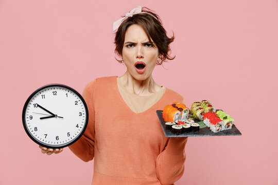 Young angry displeased disturbed disssatisfied woman 20s wear sweater hold in hand makizushi sushi roll served on black plate traditional japanese food clock isolated on plain pastel pink background