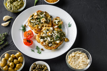 Cauliflower steak with spices, chimichurri sauce, almond flakes, olives, fried cherry tomatoes and capers on a white plate. Dark background.