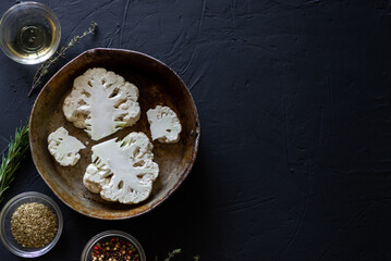 cauliflower steak cooking, recipe. Raw parts of cauliflower lie in a frying pan. Near olive oil, spices. Dark background. Place for text.