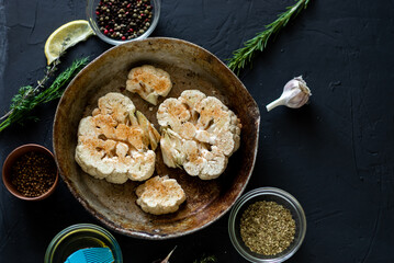 Cauliflower steak cooking. Raw cauliflower sprinkled with spices lies in a frying pan. Olive oil, herbs, various spices nearby. Dark background.