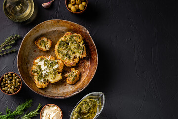 Cauliflower steak with spices lies in a frying pan. Olive oil, chimichurri sauce, capers, olives, herbs, various spices side by side. Dark background. Vegetarian food.