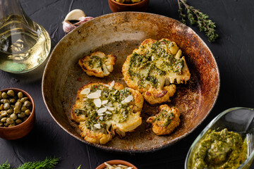 Cauliflower steak with spices lies in a frying pan. Olive oil, chimichurri sauce, capers, olives, herbs, various spices side by side. Dark background. Vegetarian food.