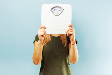 Woman with weight scale. Young slim woman hiding her face behind weight scale on blue background