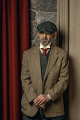 An adult man with a cane is dressed in the style of Peaky Blinders, looking away