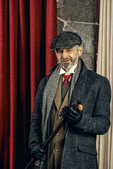 An adult man with a cane is dressed in the style of Peaky Blinders, looking away