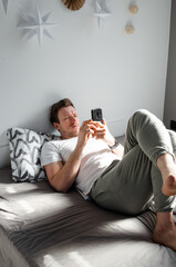 Caucasian attractive young man in white t-shirt woke up chatting, surfing, working on pc and making phone call on bed in bedroom interior. Modern device for remote communication