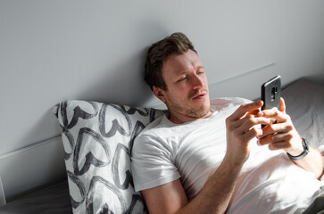 Caucasian attractive young man in white t-shirt woke up chatting, surfing, working on pc and making phone call on bed in bedroom interior. Modern device for remote communication