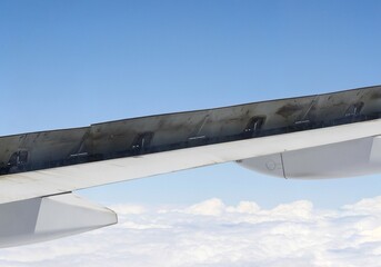 View of above clouds and wing of airplane with open flaps from window with blue sky background. Use for wallpaper or backdrop.