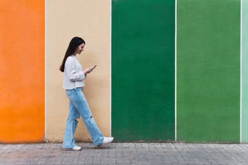 Asian woman walking while using her smartphone against colorful wall - Powered by Adobe