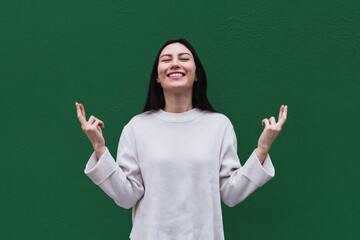 Asian woman smiles broadly with eyes closed, keeps fingers crossed, hope for good luck during exam, isolated over green background with copy space for your text