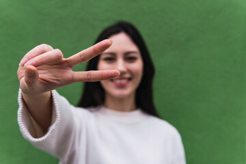 Fototapeta premium Portrait of happy asian girl stands against green wall background, shows hand gesture of peace, looks into camera and smiles. Focus on fingers