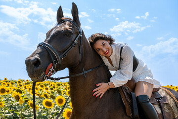 Obraz premium Cheerful brunette horseback riding. She is wearing traditional white dress and long black boots. Rural scene. Sunny day. Sunflower field in background.