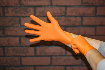 The hands of a man wearing rubber gloves are insulation against a brick wall. Close-up of orange latex protective gloves against a brick wall.