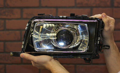 A man holds a headlight removed from a car during repairs against a brick wall. Repair and tuning...