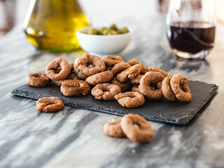 Taralli with red wine on a black slate plate, on a marble table. Vegan aperitif. Red wine, olive oil and olives in the background