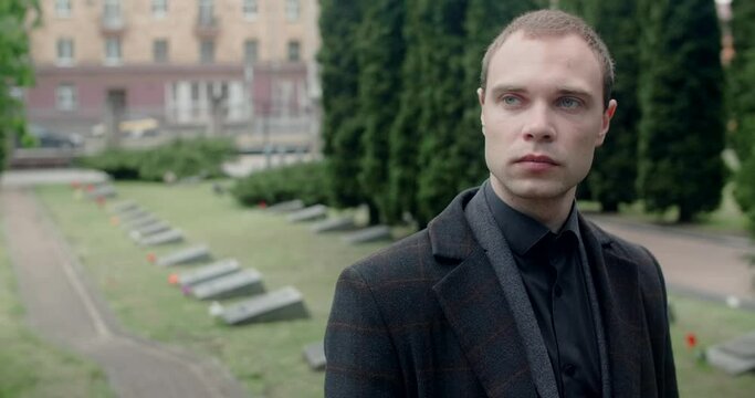 Sad young man in suit and overcoat stands on the cemetery. Funeral and farewell, honoring the passed relatives, 4k 60p Prores