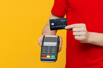 Cropped delivery guy employee man wear red T-shirt uniform work as dealer courier hold wireless bank payment terminal customer process acquire credit card payment isolated on plain yellow background.