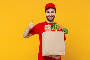 Smiling delivery guy employee man in red cap T-shirt vest uniform workwear work as dealer courier hold craft brown paper bag with grocery food show thumb up isolated on plain yellow background studio