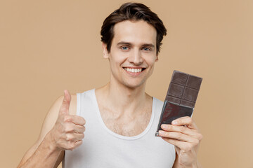 Attractive young man 20s perfect skin in undershirt hold dark chocolate bar show thumb up isolated...