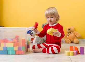 A beautiful little child in red pajamas is sitting on floor in playroom and playing with colorful cubes of figures folded in transparent box